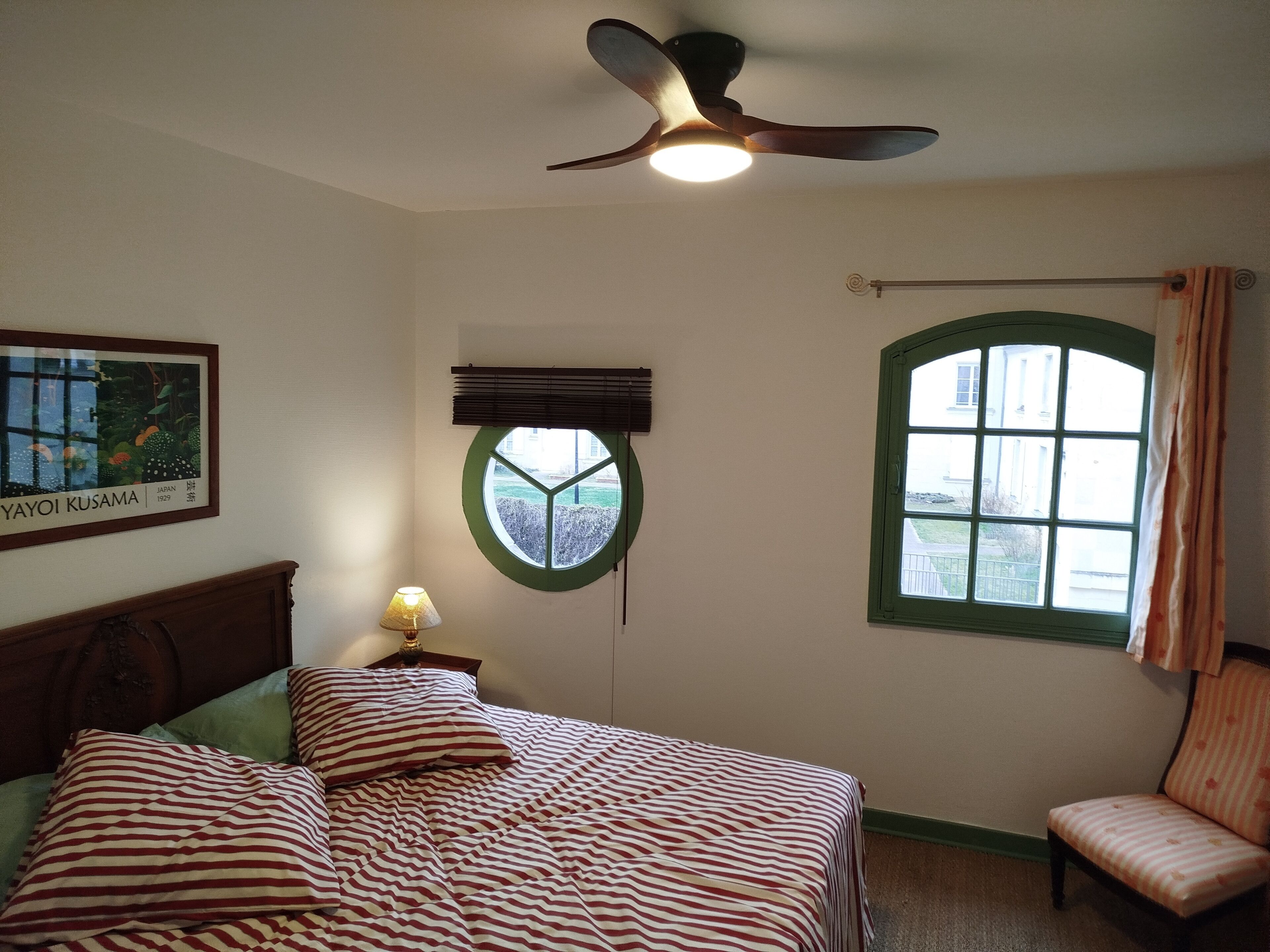 Quarto com uma cama, uma cadeira, uma janela, um ventilador de teto e uma luminária fixada na parede.