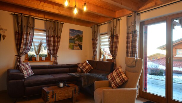 Ein gemütliches Wohnzimmer mit einem braunen Sofa, einem beige farbenen Sessel und Karottenvorhängen. Drei Pendelleuchten sind angebracht und durch das Fenster ist ein Haus zu sehen.