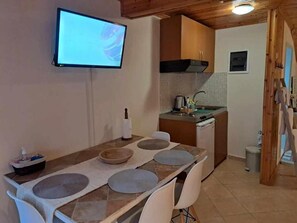 A compact kitchen with a TV mounted on the wall, a dining table with placemats, and a small sink. - 🏖Sunny Family Studio • 60m Walk to Beach • Parking & Garden (Skala Marion)