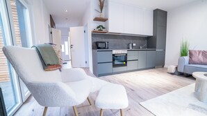 Een moderne woonkamer met een grijze bank en een poef, een houten vloer, en een keuken met grijze kastjes en een magnetron.