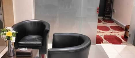 Uma área de recepção com duas poltronas de couro preto, uma mesinha com um vaso de flores e um tapete vermelho que leva a uma porta com um adesivo em silhueta de mesquita.