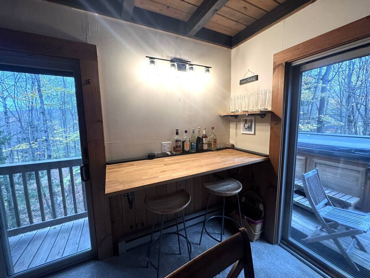 A cozy bar area with a wooden counter, two stools, and a view of the outdoors through a large window.