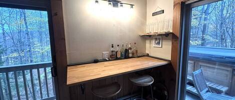 A cozy bar area with a wooden counter, two stools, and a view of the outdoors through a large window.