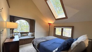Un dormitorio con tragaluz, cama con ropa de cama azul y blanca, escritorio con lámpara y asiento junto a la ventana con vista a árboles.