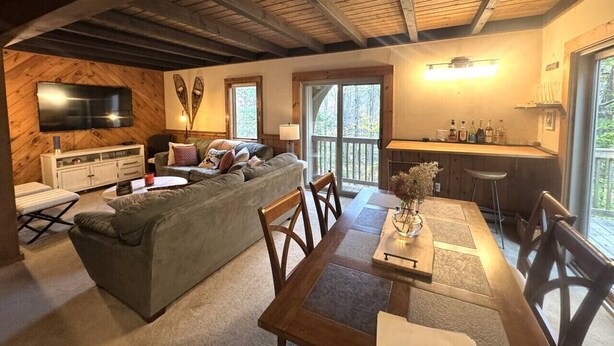 Un salón acogedor con sofá, mesa de comedor y sillas, zona de cocina con barra, y grandes ventanales que ofrecen vistas al exterior.