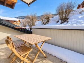Een houten tafel en stoelen op een balkon, met sneeuw op de grond en een uitzicht op huizen en bomen in de verte.