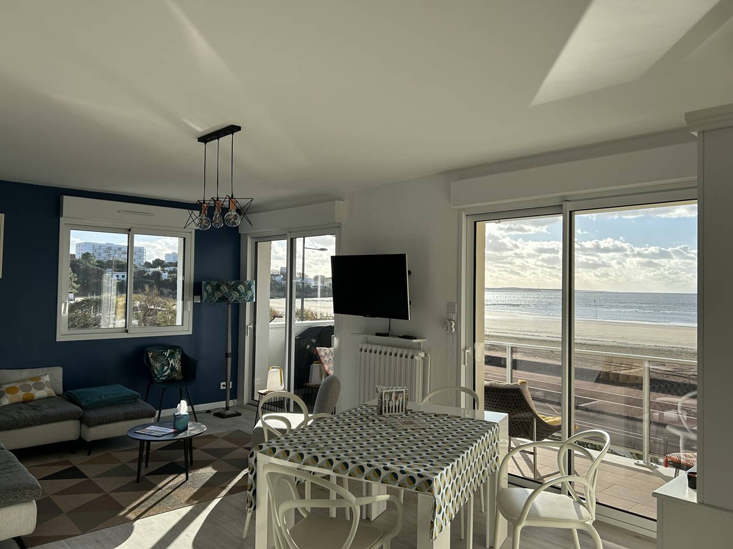 Ein modernes Wohnzimmer mit großem Fenster mit Blick auf einen Strand, einem Essbereich mit weißen Stühlen und einem an der Wand befestigten Flachbildfernseher.