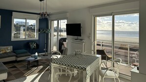 A modern living room with a large window overlooking a beach, a dining area with white chairs, and a flat-screen TV mounted on the wall. - Apartment T4 - 6 ppl - Parking - Sea View in St Georges de Didonne (ST GEORGES DE DIDONNE)