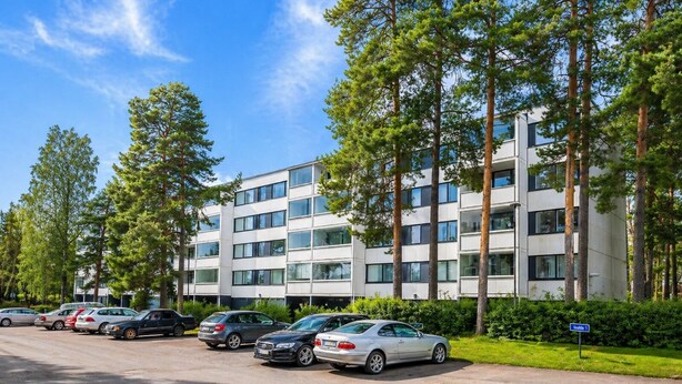 Ein mehrstöckiges Gebäude mit Parkplatz davor, umgeben von Bäumen.