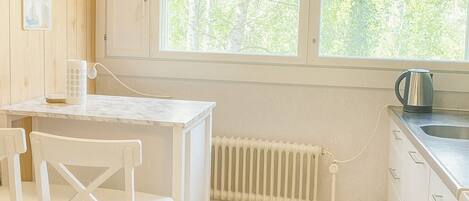 A compact kitchen with a wooden floor, white cabinetry, a white table with chairs, and a window overlooking greenery.