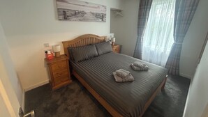 Een slaapkamer met een houten bed, een nachtkastje met een lamp, een schilderij aan de muur en een raam met gordijnen.