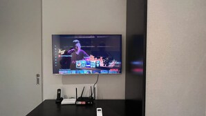 A flat-screen TV mounted on a wall displaying a show, a black desk with a router, a phone, and a small speaker. - SCARLETZ SUITES KLCC BY LLP (Kuala Lumpur)