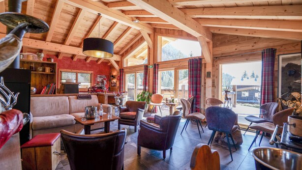 L’intérieur chaleureux d’un chalet en rondins, avec un plafond en bois, un canapé, plusieurs fauteuils, une table avec des boissons et une vue sur l’extérieur.