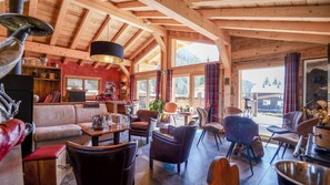 Een gezellig chaletinterieur met houten plafond, een bank, meerdere stoelen, een tafel met drankjes, en uitzicht op de omgeving.