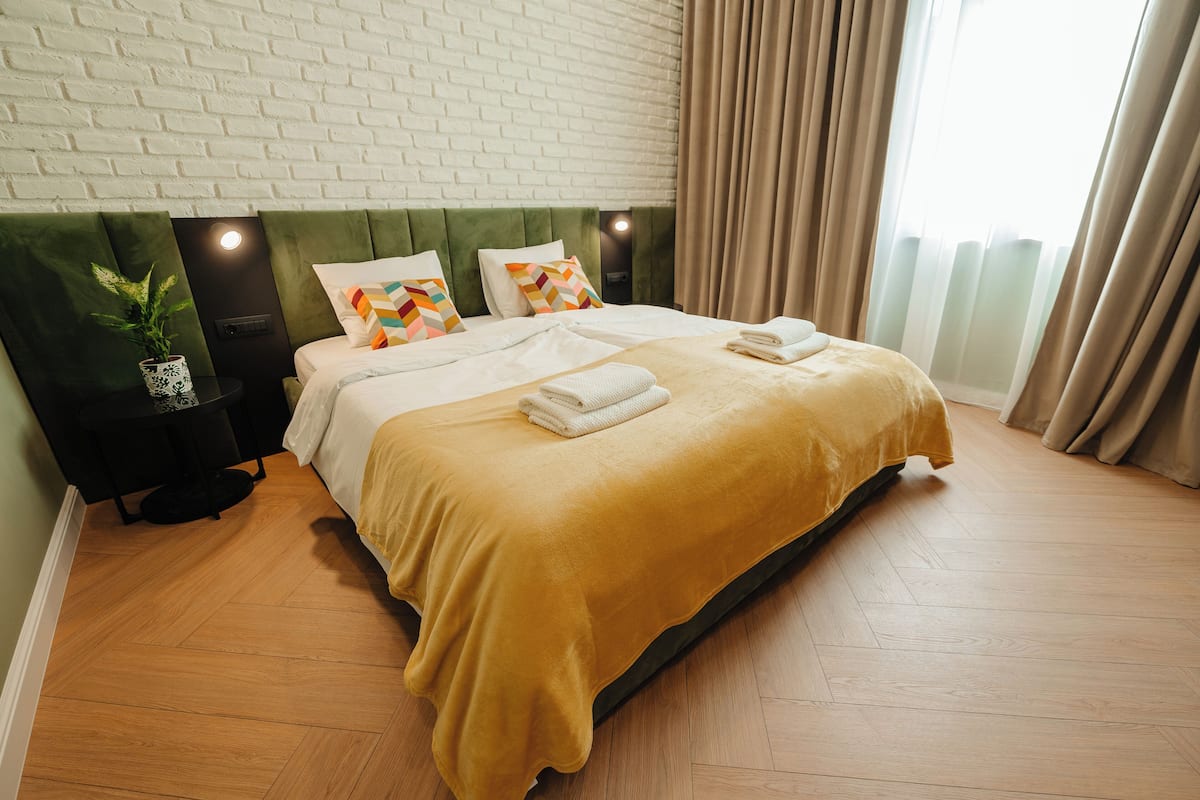 A modern hotel room with a brick wall, a green upholstered headboard, a large bed with a yellow bedspread, two bedside tables with lamps, and a potted plant.