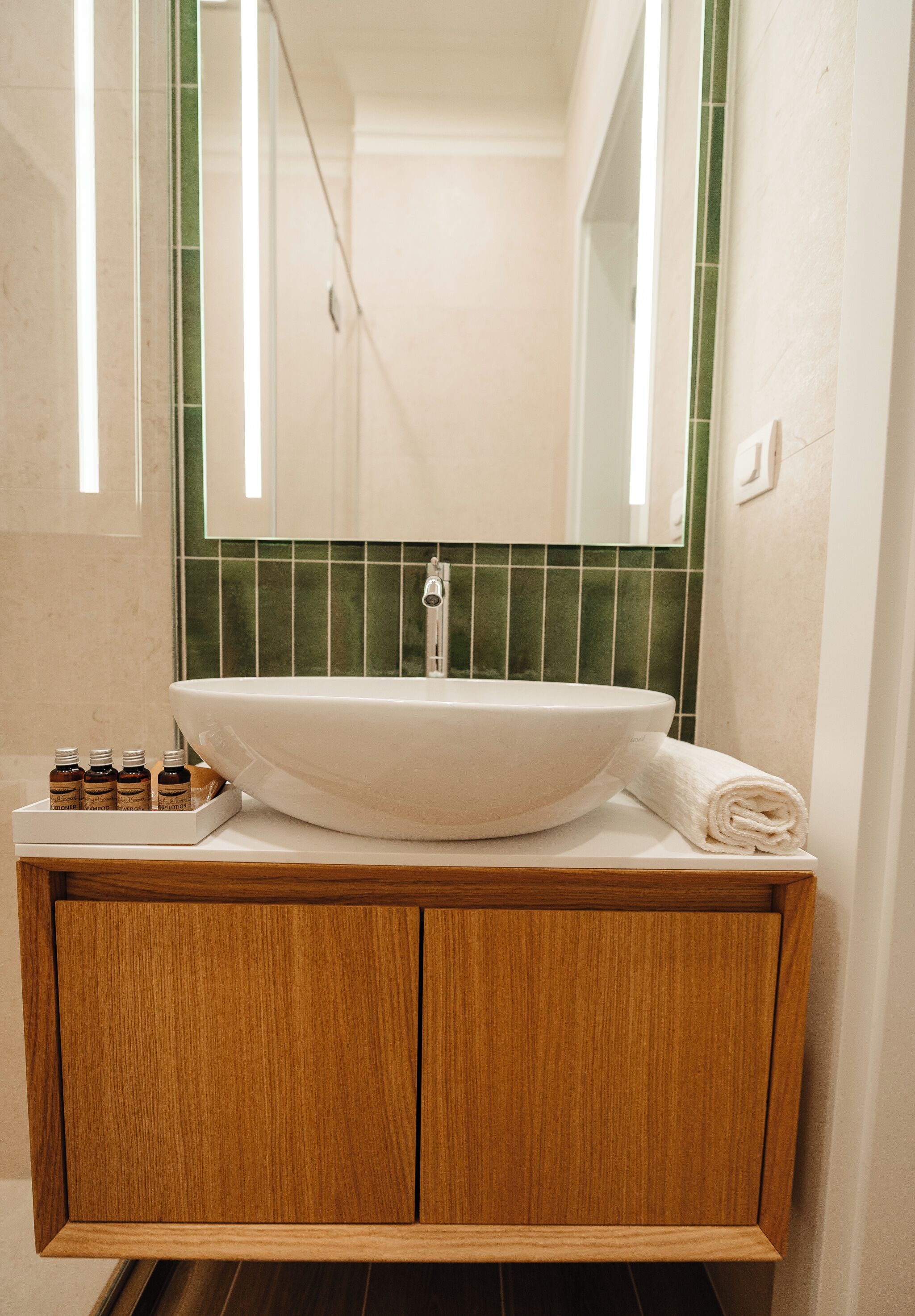 Bagno con lavabo bianco, mobile contenitore in legno, schienale rivestito in piastrelle verdi e uno specchio sopra il lavabo.