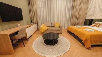A modern hotel room with a flat-screen TV, a desk with a chair, a round coffee table, a sofa with cushions, a bed with a yellow blanket, and a wall-mounted TV.