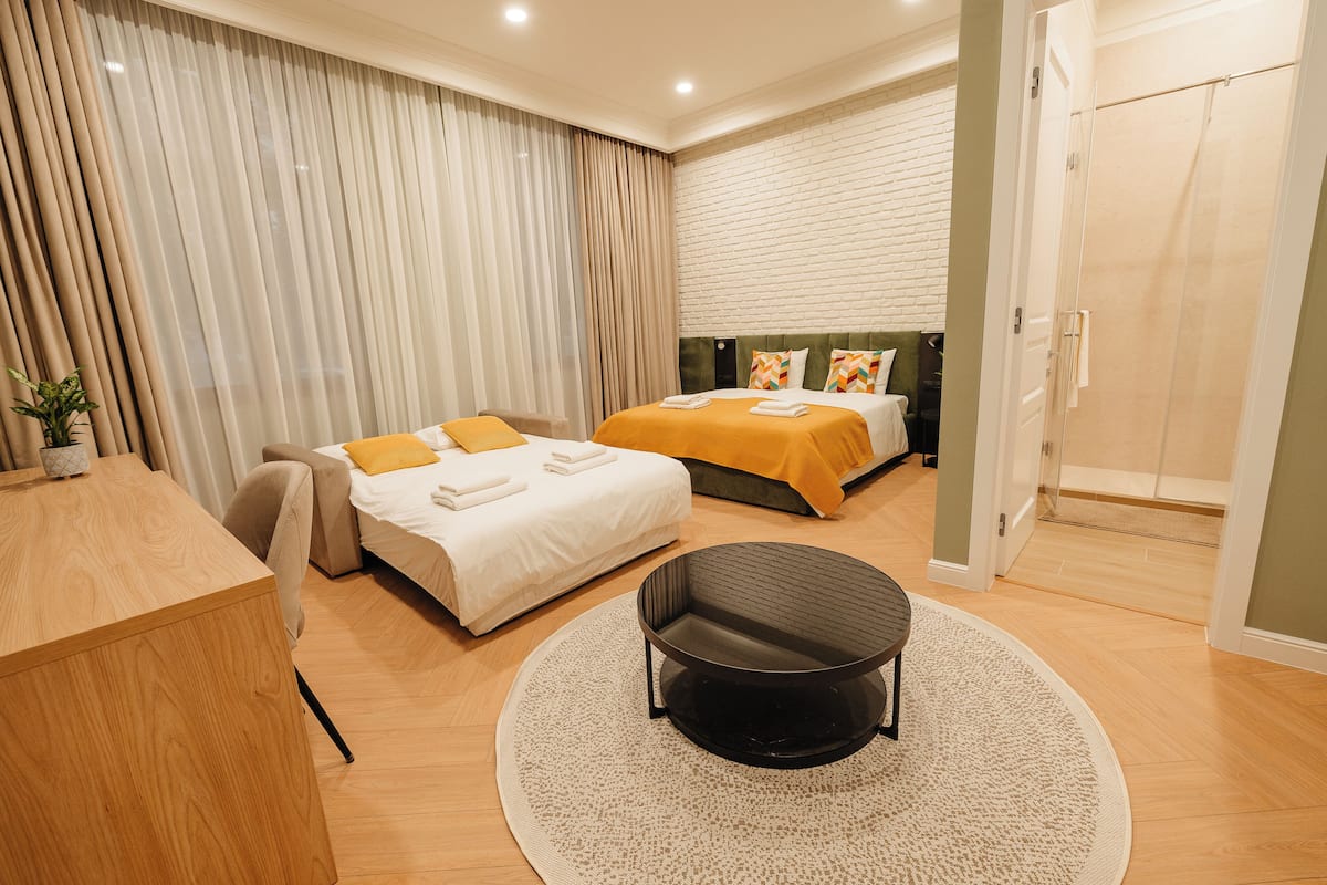 A modern hotel room with two beds, a desk, a round table, and a large window with curtains.