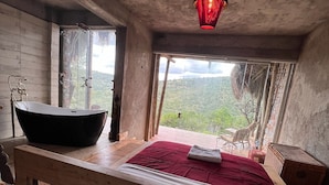 Ein hölzernes Bett mit roter Decke, eine schwarze Badewanne und eine rote Pendelleuchte in einem Zimmer mit großen Fenstern.
