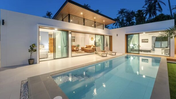 Une maison moderne dotée d’une piscine, de grandes portes vitrées donnant sur un espace de vie aménagé, et offrant une vue sur des palmiers.
