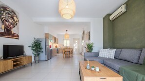 A modern living room with a grey sofa, a wooden coffee table, and a TV on a wooden stand. There is a kitchen in the background with a dining area. - Villa Nero -
A beautiful 2-bedroom villa with Mini Pool, 5 min from the beach! (PLAKIAS)