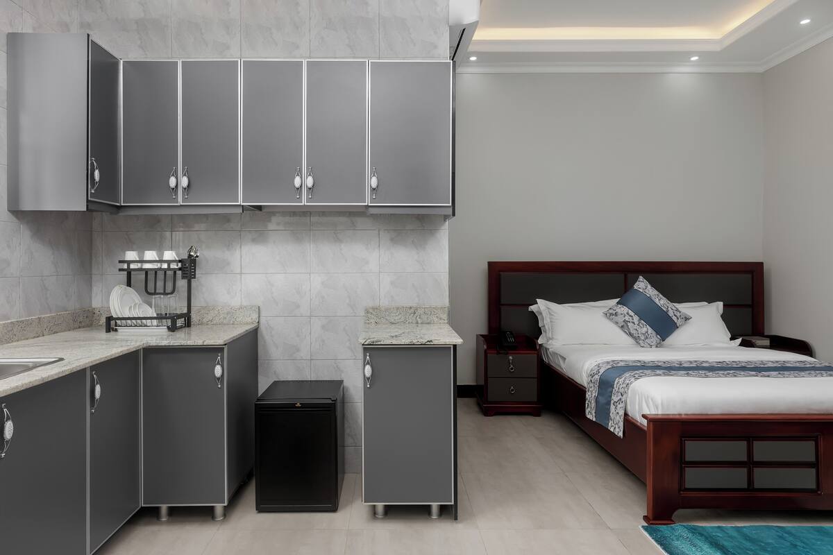 A modern hotel room with a kitchen area, a bed with a wooden headboard, and a small bedside table.