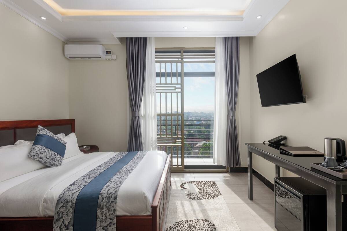 A hotel room with a bed, a desk, a flat-screen TV, and a view through a large window.