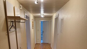 Un couloir doté d’un plancher en bois, de murs blancs et d’un plafond avec des poutres en bois. On y trouve des crochets pour manteaux sur le mur de gauche et une petite échelle en bois posée sur le sol.