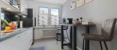 A modern kitchen with white cabinets, a black countertop, and a window with a view. There is a chair and a table with a vase and a book on it.
