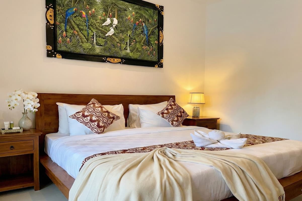 A neatly made bed with white linens and patterned pillows, a wooden headboard, a nightstand with a vase of white flowers, and a painting of birds on the wall.