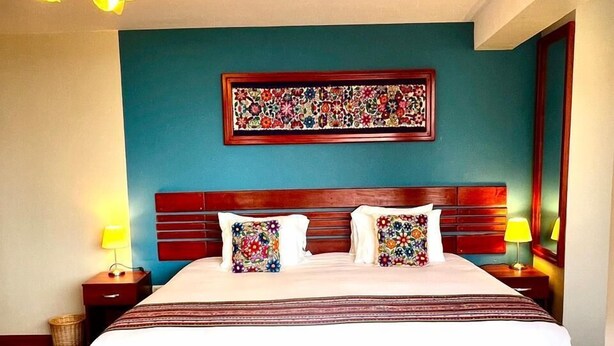 A hotel room with a large bed, wooden headboard, two bedside tables with lamps, a wall-mounted artwork, and a wooden floor. - Hotel Chinchero Boutique & Tours (Chinchero)