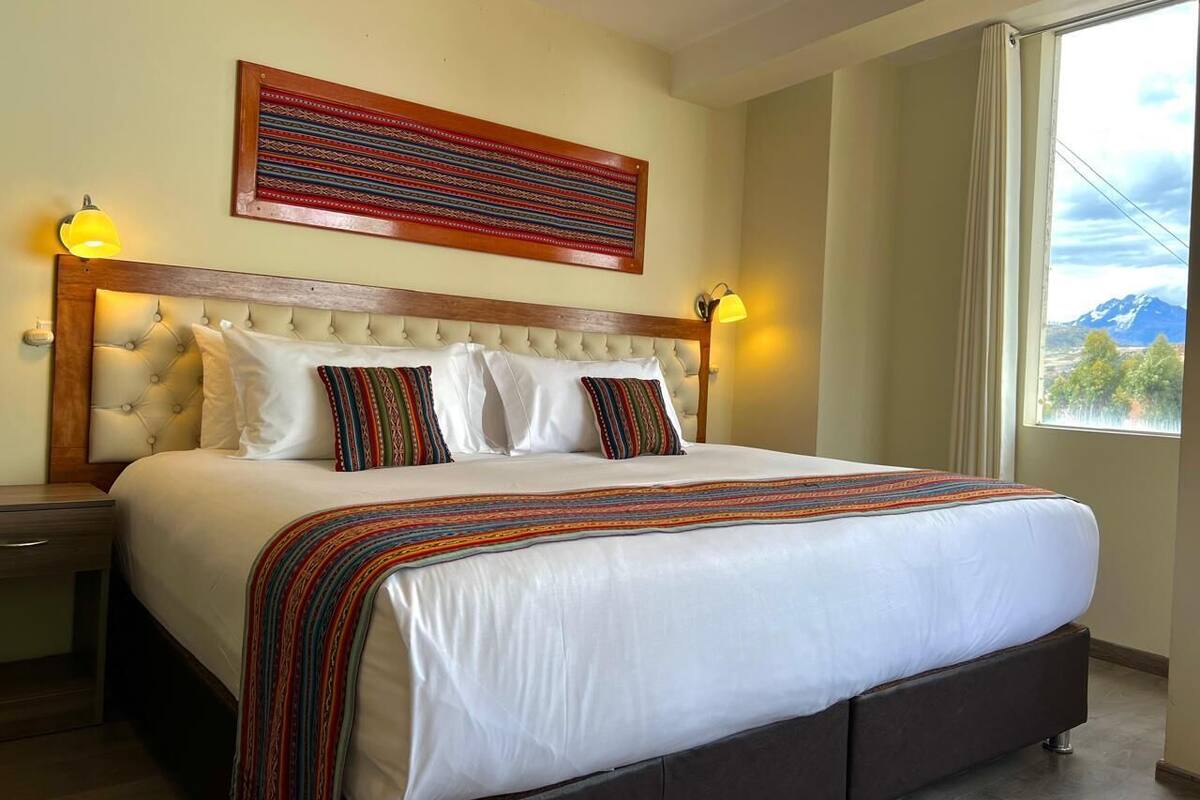 A large bed with a tufted headboard, white bedding, and colorful pillows. There are two bedside lamps and a large window with a mountain view.