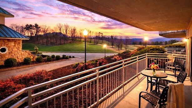 A balcony with outdoor seating, a stone building, and a view of a golf course at sunset. - Golf Course Views • Fireplace • Free DOLLYwood Ticket • Near Dollywood (Pigeon Forge)