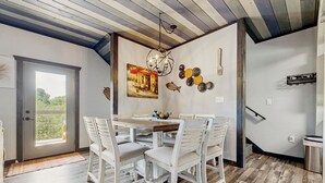 Un comedor con una mesa de madera y sillas blancas, un cuadro en la pared y vistas a través de una puerta.