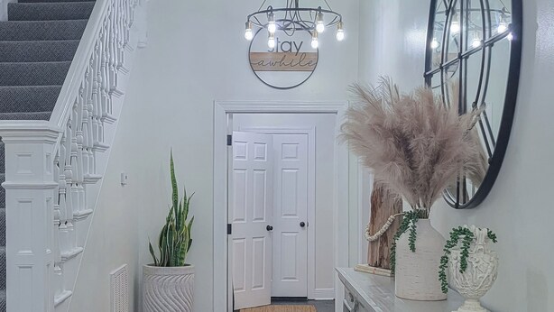 A staircase with white balusters, a white door, a wall-mounted sign with lights, and a mirror with a black frame. - 4 BR Home w/ Modern Comforts & Park View (Savannah)