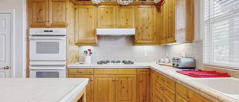 Una cocina con gabinetes de madera, una encimera blanca, horno empotrado, fregadero y una ventana con persianas.