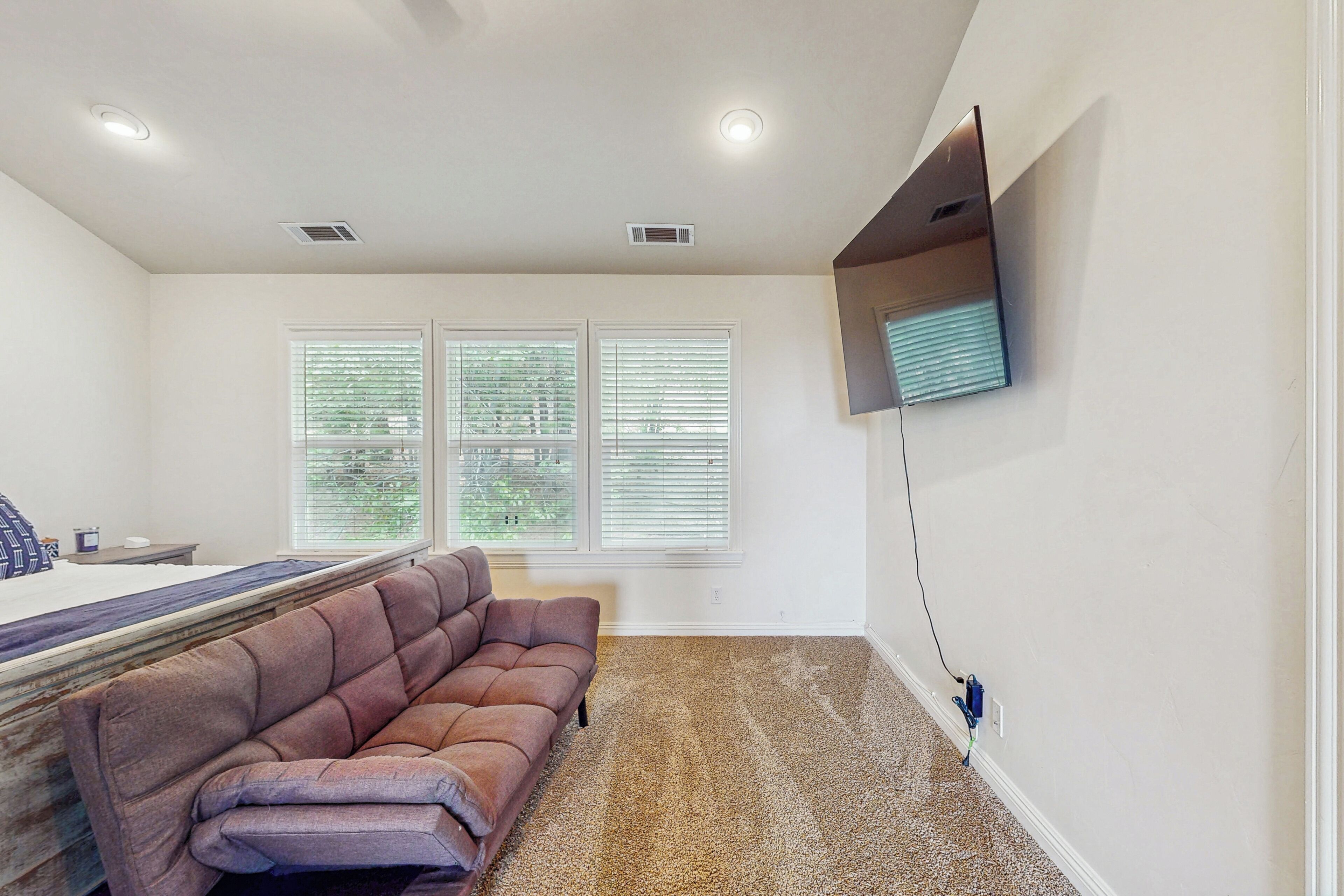 A bedroom with a bed, a brown sofa, a TV mounted on the wall, and a window with blinds.