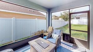 Une terrasse ensoleillée avec un fauteuil en osier, une petite table et une vue sur un jardin.