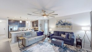 Un salon moderne comprenant un canapé, un pouf et une table basse, un coin cuisine avec des placards et des appareils électroménagers, et un ventilateur de plafond.