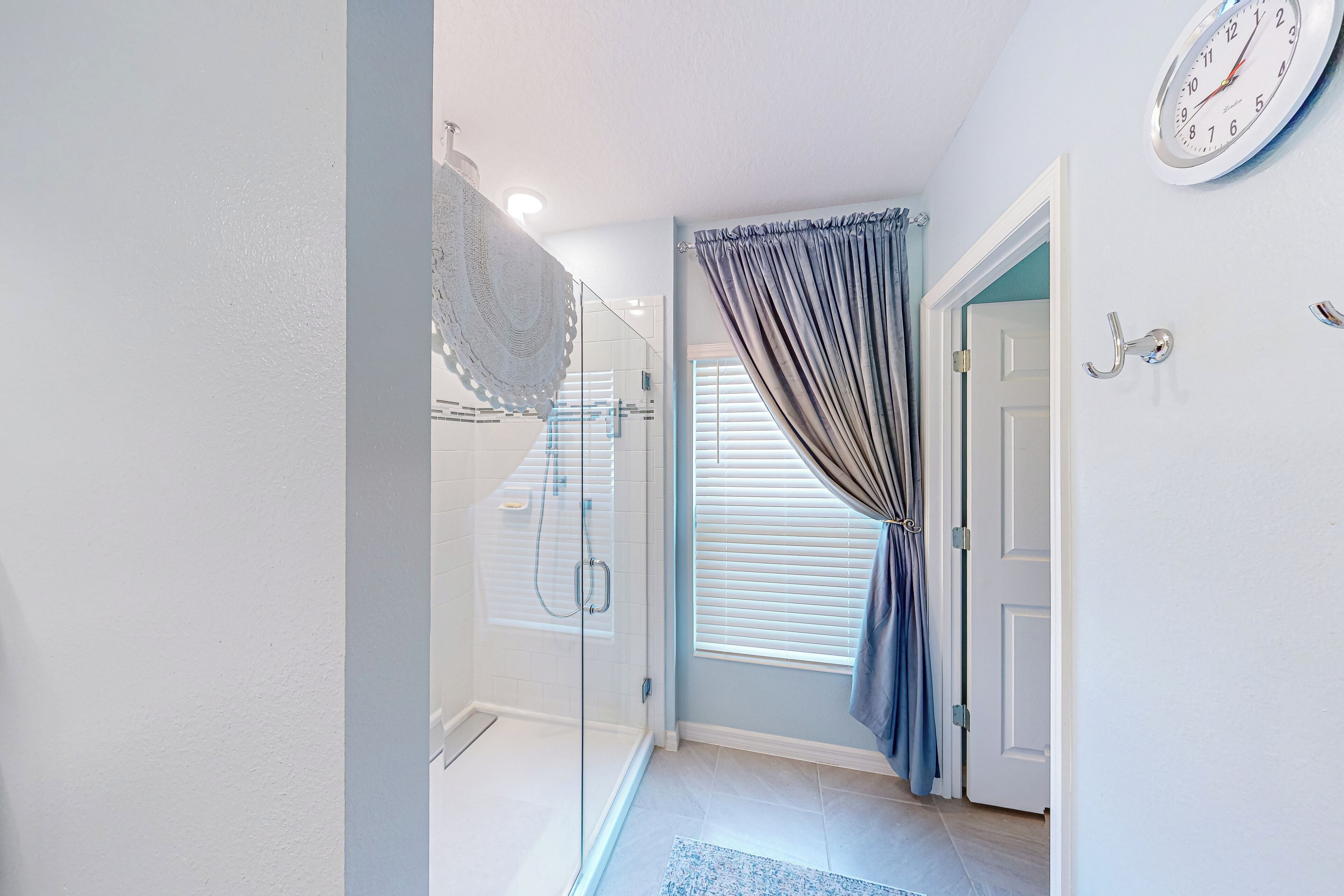 Une salle de bain avec une douche à parois de verre, un porte-serviettes, une horloge au mur et une porte donnant sur une autre pièce.