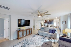 A spacious living room with a large flat-screen TV, a ceiling fan, and a modern kitchen in the background. - 3BR House w/ Enclosed Yard & Gas Grill (Groveland)