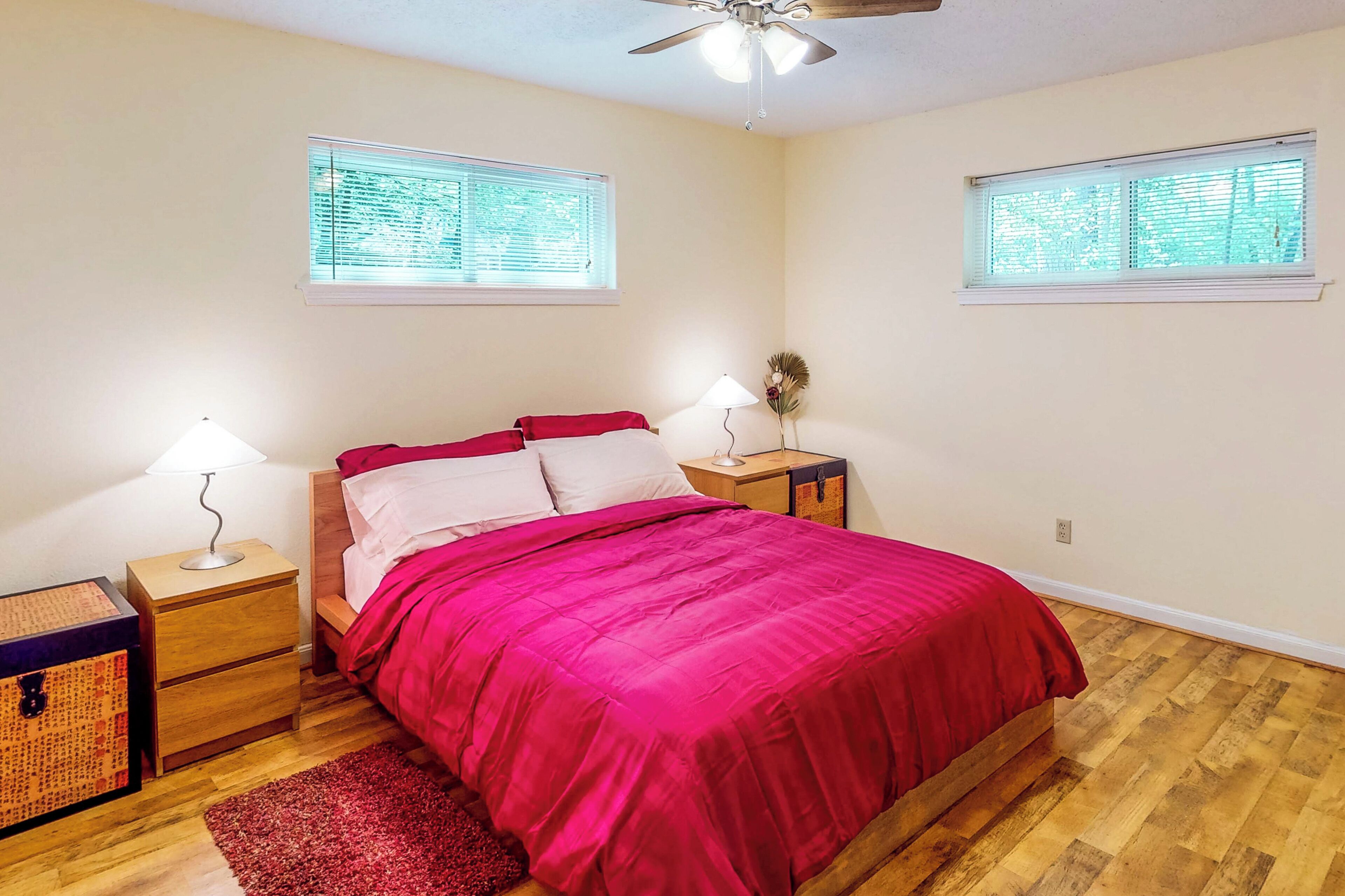 A bedroom with a wooden floor, a bed with red bedding, two bedside tables with lamps, and two windows with blinds.