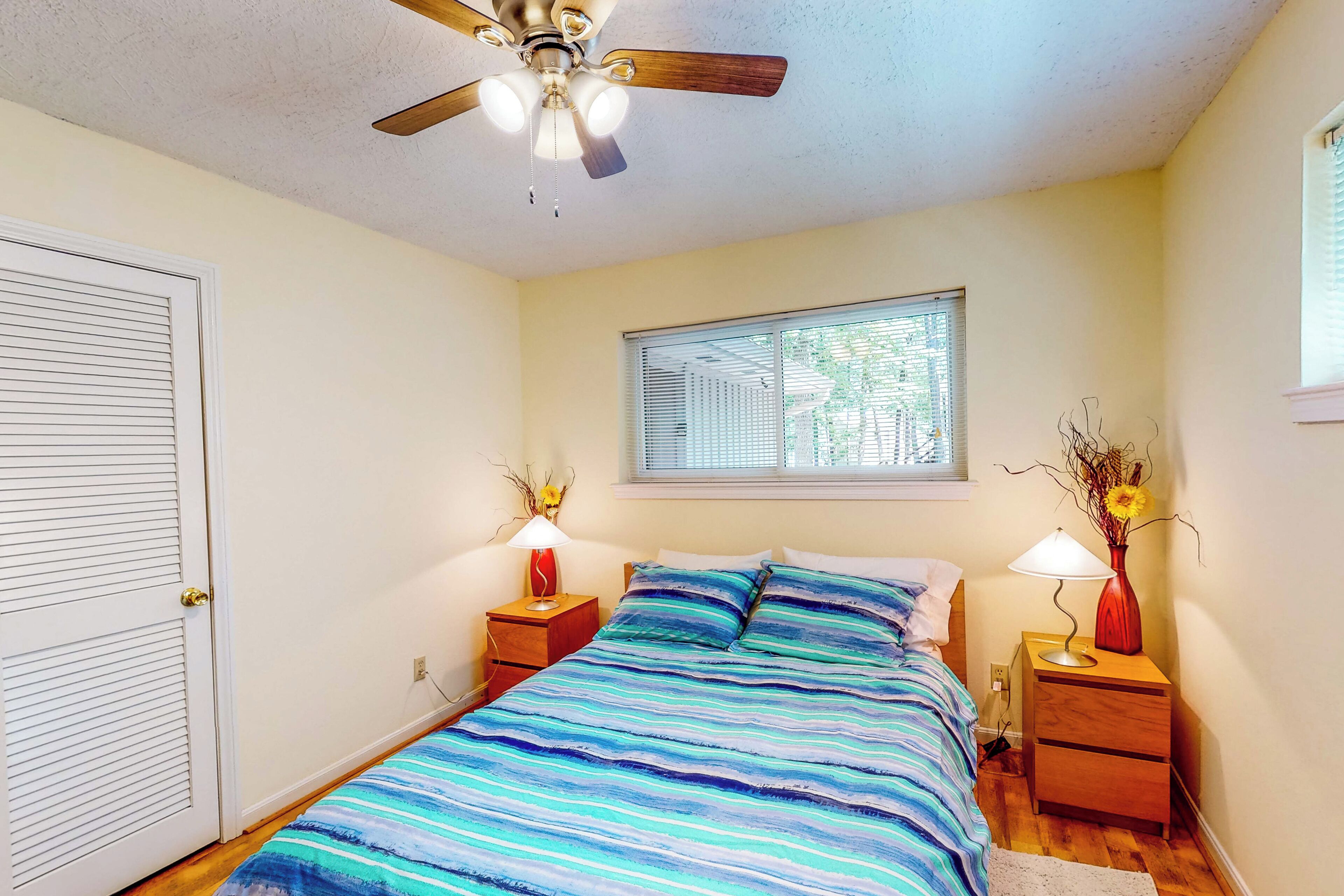 A bedroom with a bed, a ceiling fan, a window, and a nightstand with a lamp.
