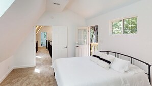 Ein Schlafzimmer mit einem großen Bett, einem Fenster mit Blick auf Bäume, einer Tür zu einem anderen Raum und einer Deckenleuchte.