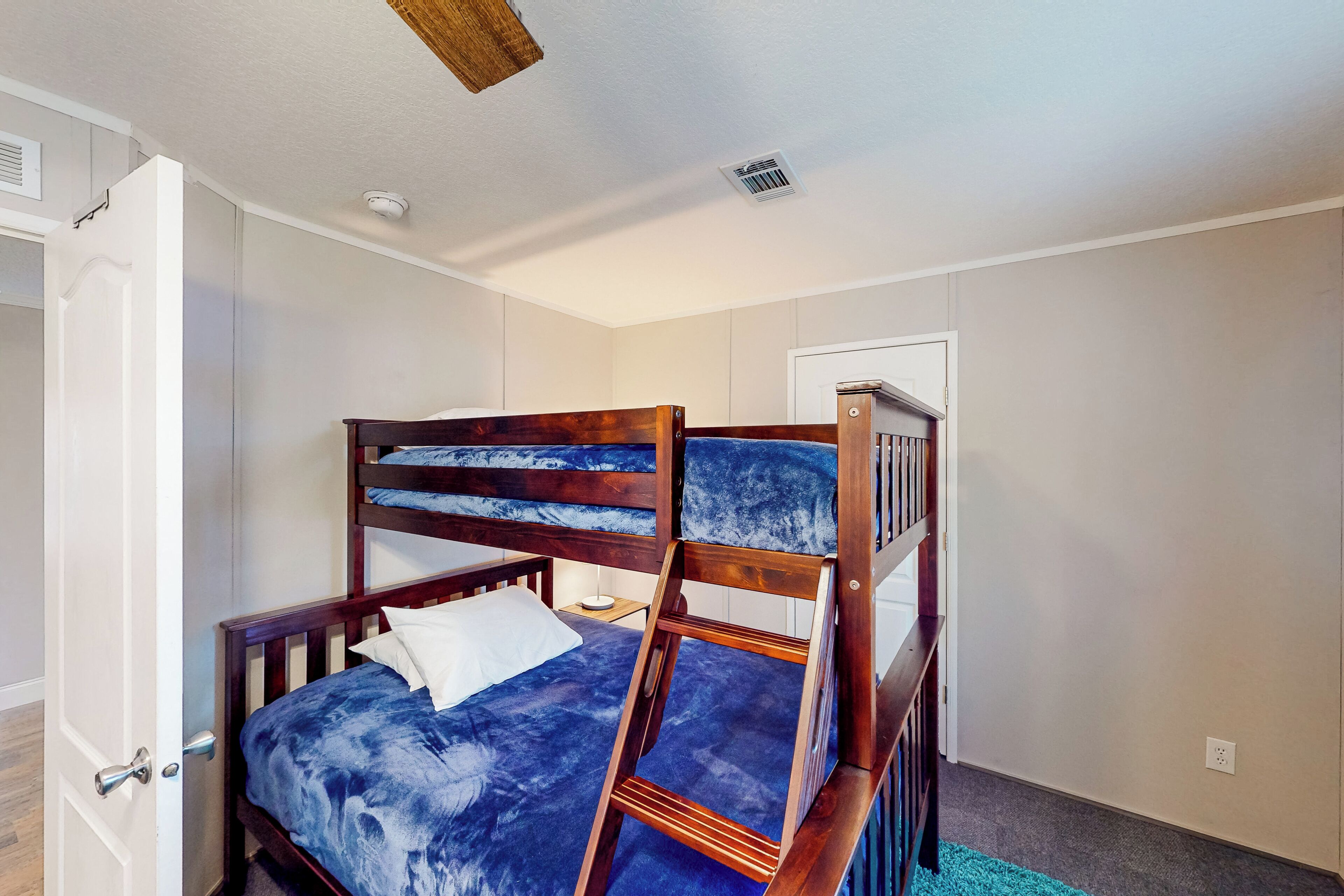 A room with a bunk bed, a door, a small bedside table, and a ceiling vent.