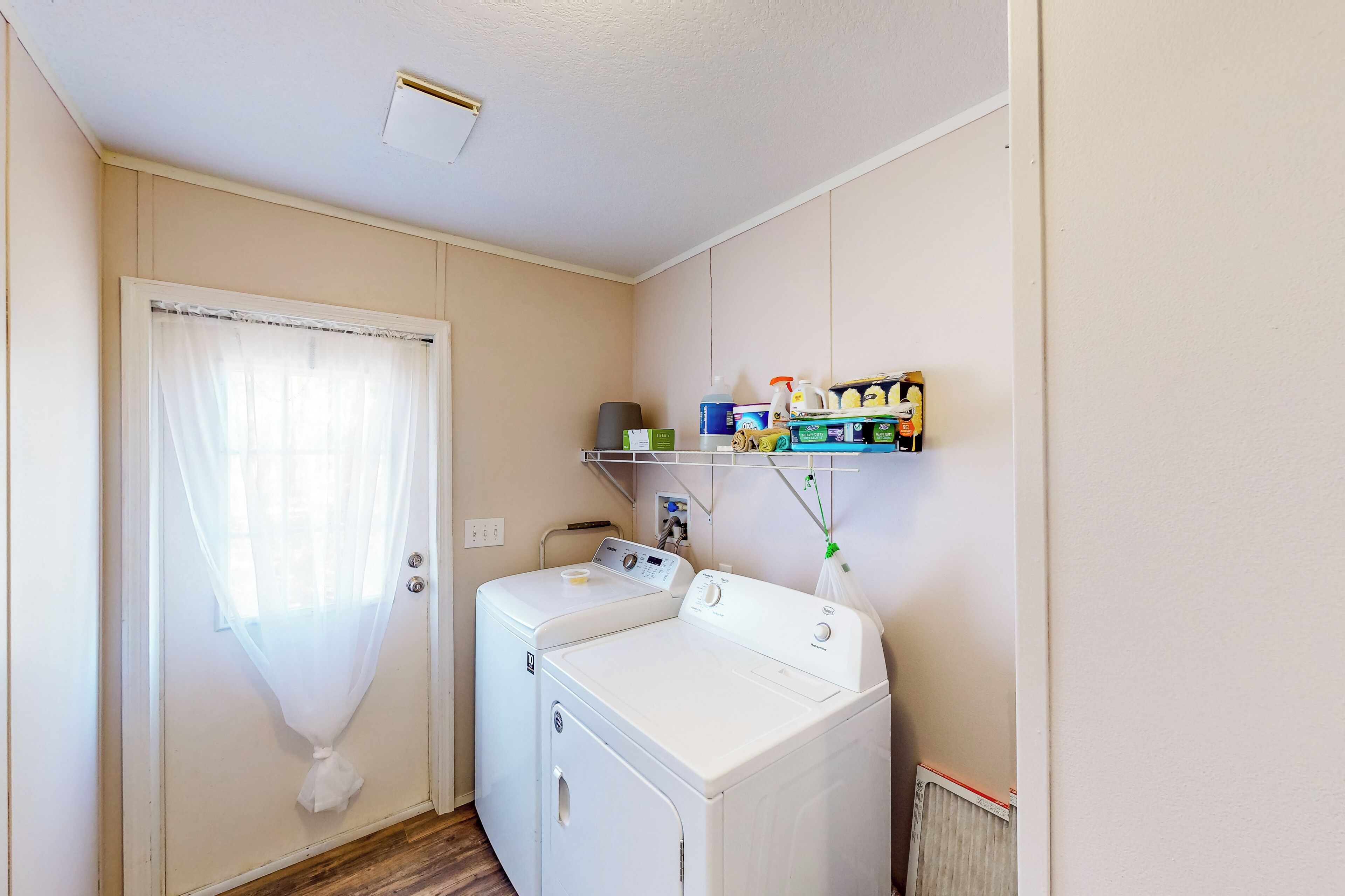 Une buanderie équipée d’un lave-linge et d’un sèche-linge, d’une étagère contenant des produits de nettoyage et d’une porte ornée d’un rideau léger.