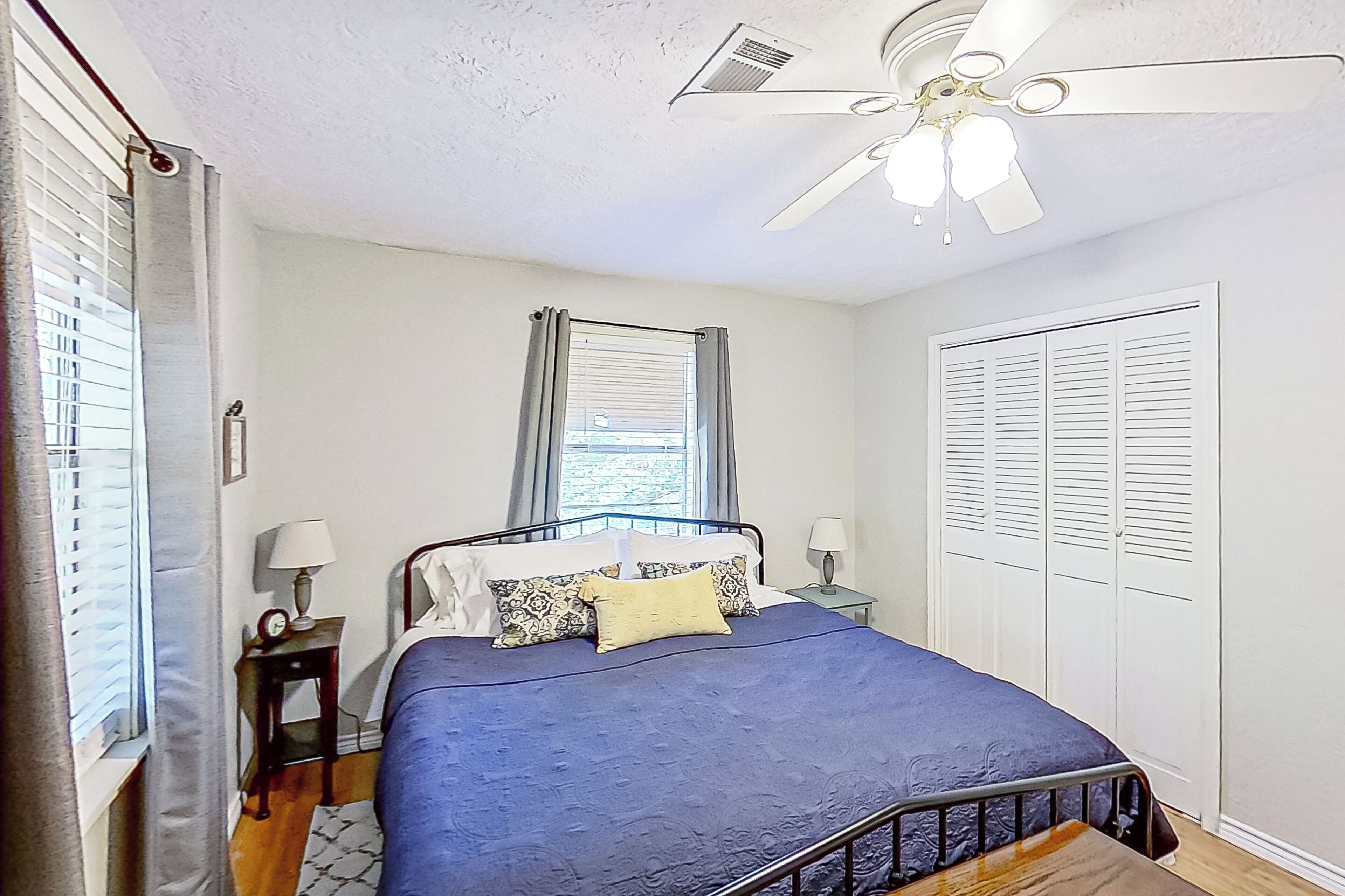 A bedroom with a bed, a ceiling fan, a window with curtains, a nightstand with a lamp, and a wardrobe.