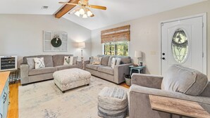 A cozy living room with a grey sofa set, a wooden ceiling fan, a large window with blinds, and a decorative wreath on the wall. - 2BR Dog-friendly Home Near Lake w/ Fire Pit (San Augustine)