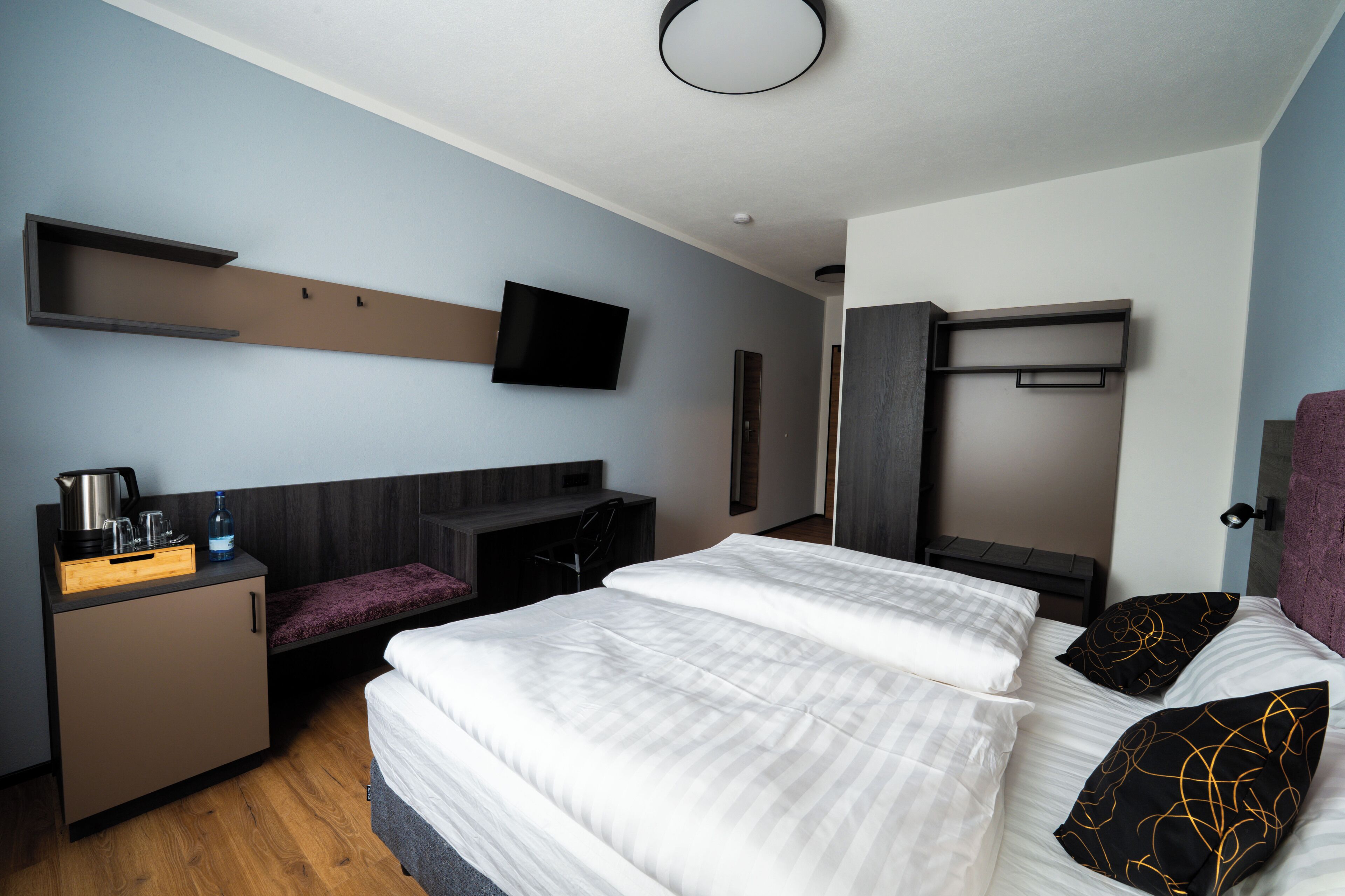 A modern hotel room with a bed, a desk, a chair, a TV, and a wardrobe.