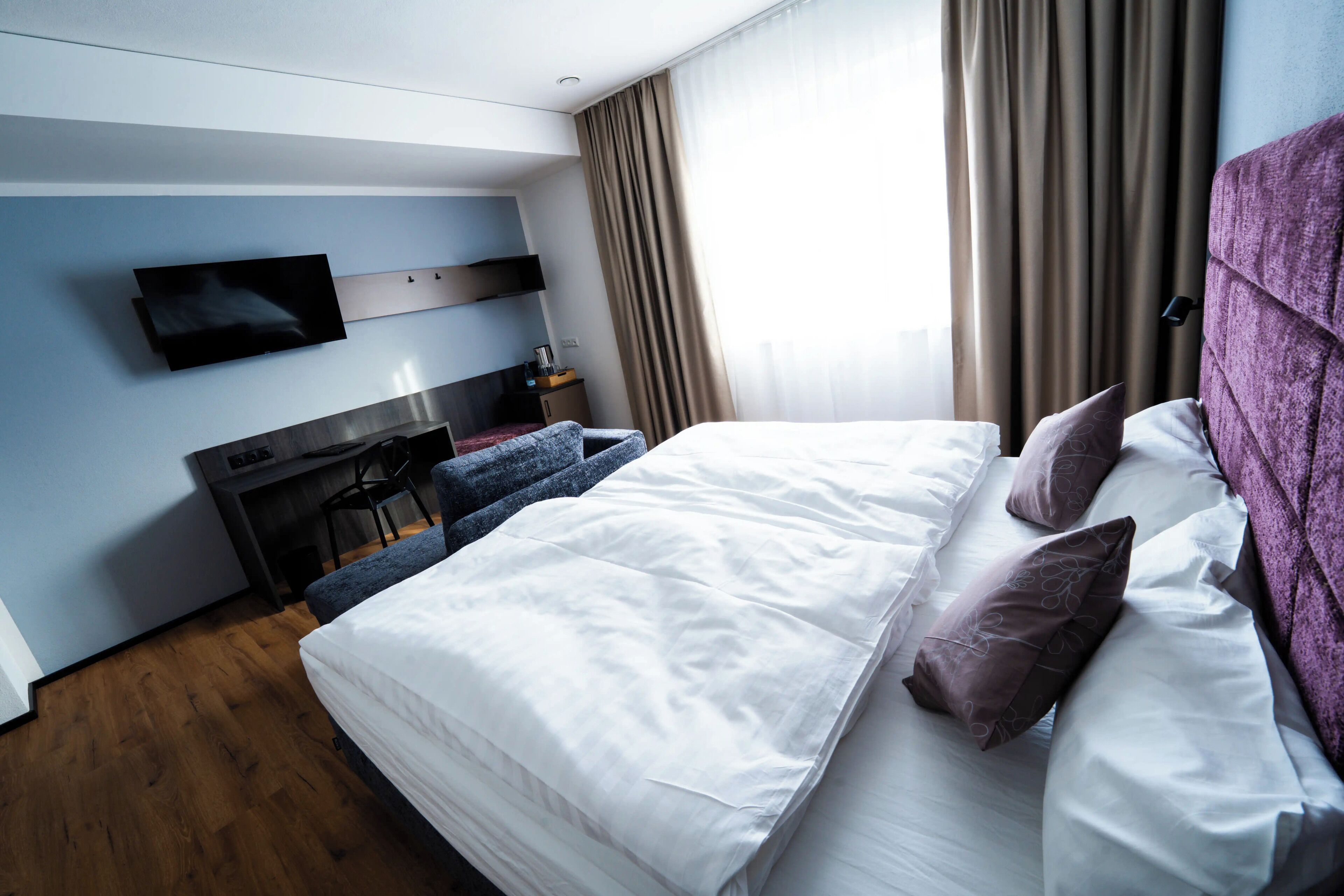 A hotel room with a bed, a desk, a flat-screen TV, and a window with curtains.
